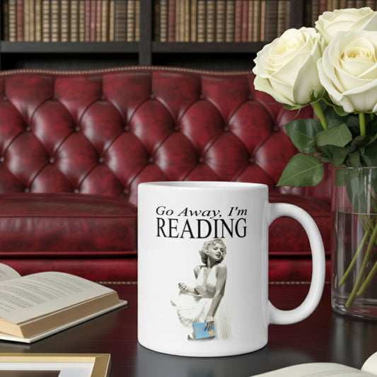 “Go Away I’m Reading” book mug on a coffee table in an elegant home library setting with a vintage red leather sofa, fresh roses, and open books. Funny gift for book lovers, readers, and literary fans who appreciate glamor and humor.
