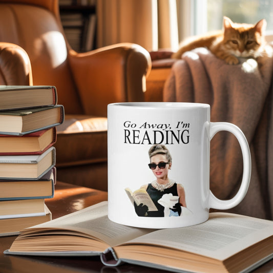 “Go Away I’m Reading” book lover mug placed on an open novel in a cozy reading nook with a leather armchair, stacked books, and a relaxed orange cat in the background — perfect gift for bookworms, readers, and introverts who love quiet time and coffee.