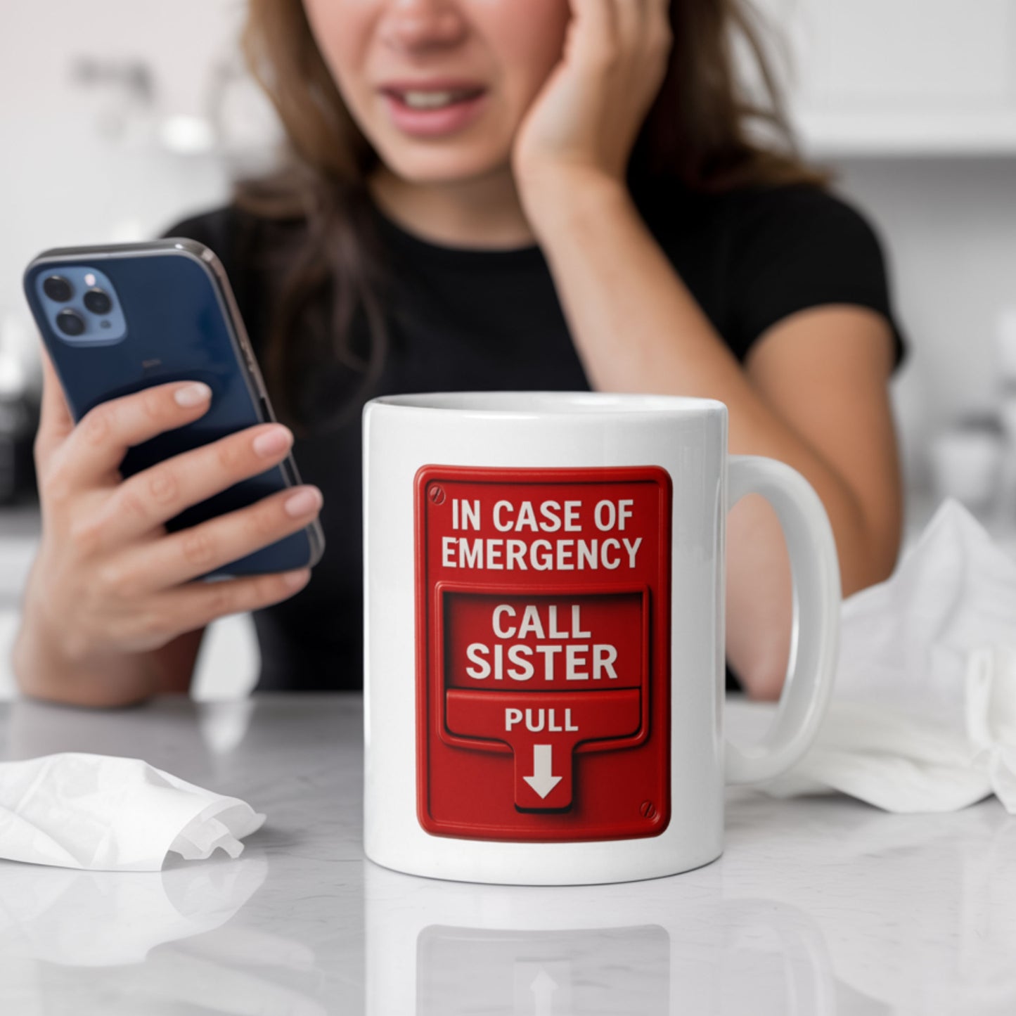 White ceramic mug with a red fire alarm-style graphic that reads “In case of emergency, call sister — pull,” pictured in a kitchen setting with a woman holding a phone and looking concerned. A funny and sentimental gift for sisters who are always the first call in a crisis.