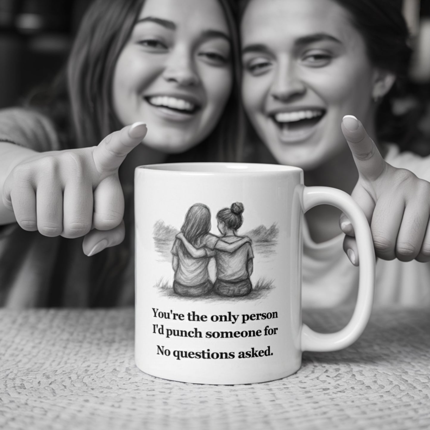 Two smiling women in cozy casual wear laugh and point at a white mug on the table that reads, 'You're the only person I'd punch someone for. No questions asked,' featuring a sketch of two girls sitting with arms around each other.