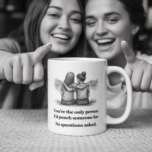 Two smiling women in cozy casual wear laugh and point at a white mug on the table that reads, 'You're the only person I'd punch someone for. No questions asked,' featuring a sketch of two girls sitting with arms around each other.