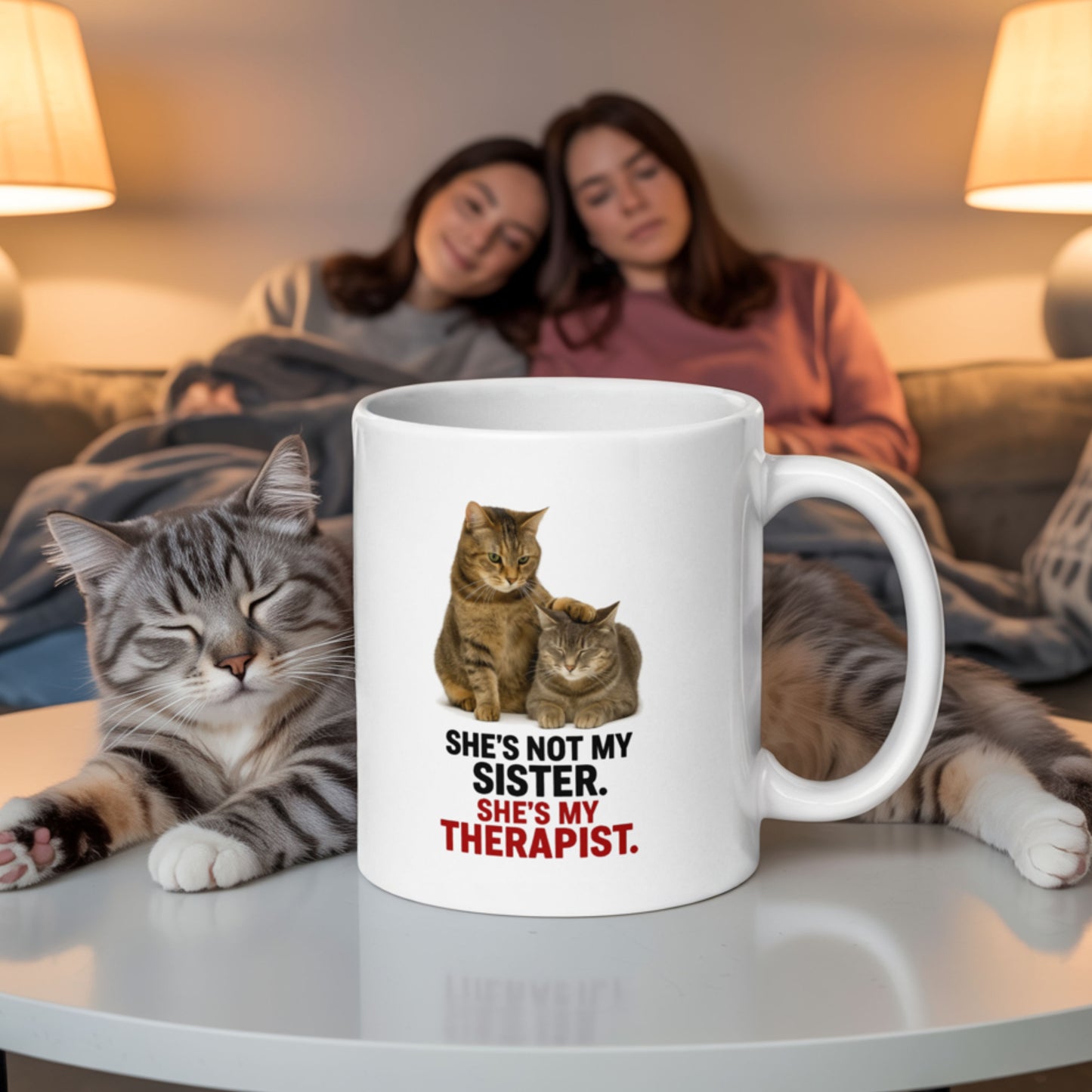 Funny sister mug that says 'She’s not my sister, she’s my therapist' featuring two cats, with one comforting the other, placed on a coffee table in front of two women relaxing on a couch and a real cat lounging nearby.