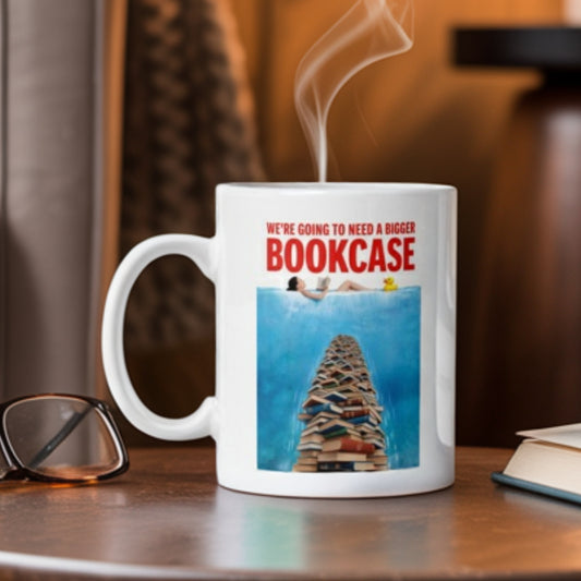 White ceramic mug with a playful design featuring a towering stack of books rising like a shark fin from the water, with a reader floating above and the caption “We’re going to need a bigger bookcase” in bold red text. A funny gift mug for book lovers, readers, and bibliophiles who never have enough shelf space.