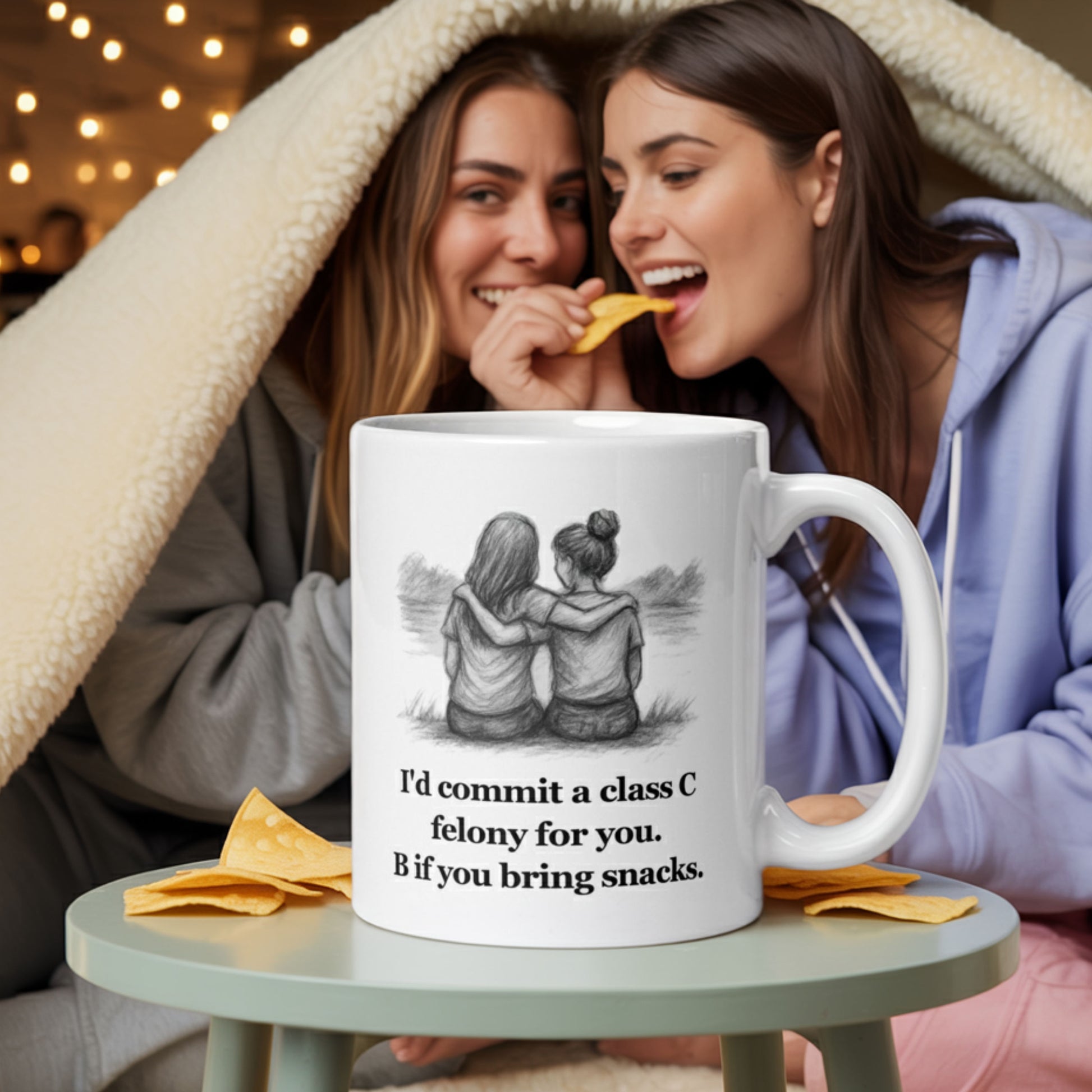 Two young women in cozy loungewear share snacks under a blanket fort, laughing together. In the foreground, a white mug with a sketch of two girls sitting side by side and the text “I’d commit a class C felony for you. B if you bring snacks.” sits on a table surrounded by chips. The scene captures playful friendship and mischief.