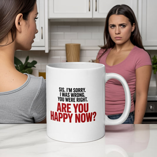 White ceramic mug with the text: 'Sis. I’m sorry. I was wrong. You were right. ARE YOU HAPPY NOW?' in bold black and red lettering, sitting on a kitchen counter. In the background, one woman looks smug while another woman in a pink top stares back with an annoyed expression, highlighting the humorous tension between sisters.