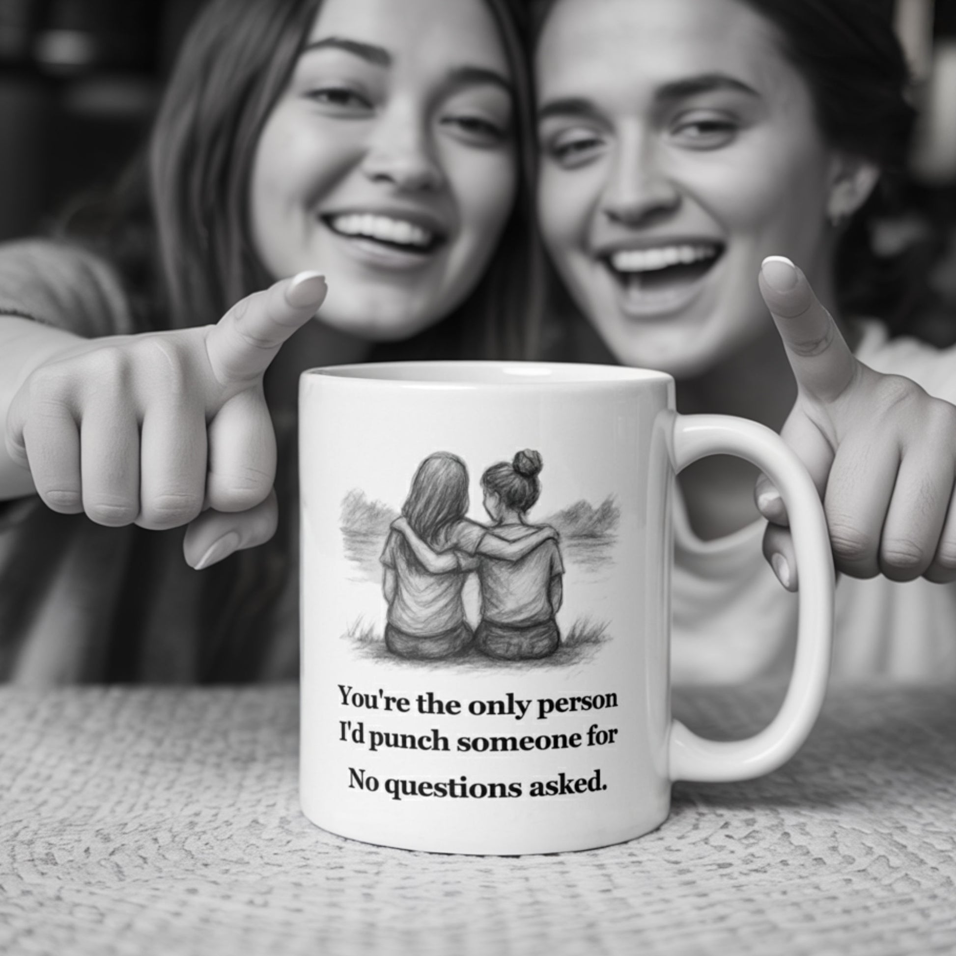 Two smiling women in cozy casual wear laugh and point at a white mug on the table that reads, 'You're the only person I'd punch someone for. No questions asked,' featuring a sketch of two girls sitting with arms around each other.