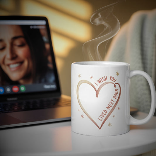 White ceramic mug with heart design and text "I Wish You Lived Next Door" sitting on a cozy table beside a laptop showing a smiling woman on a video call, with warm sunlight and steam rising from the coffee—ideal gift for long-distance friends, besties, sisters, or moms.