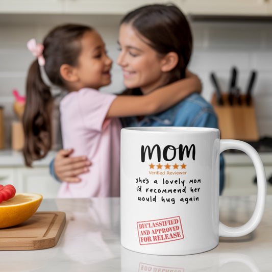 Funny 5-star review mug for mom shown in a bright kitchen setting. White ceramic coffee mug reads 'Mom – she’s a lovely mom, I’d recommend her, would hug again' with a ‘Declassified and Approved for Release’ stamp. Mom and daughter share a hug in the background. Perfect gift for mom, Mother’s Day mug, or funny mum mug from daughter or son.
