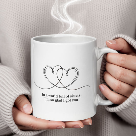 Close-up of a white ceramic mug held in cozy sweatered hands, with steam rising from the top. The mug features two intertwined hearts and the sentimental message: “In a world full of sisters, I’m so glad I got you.”
