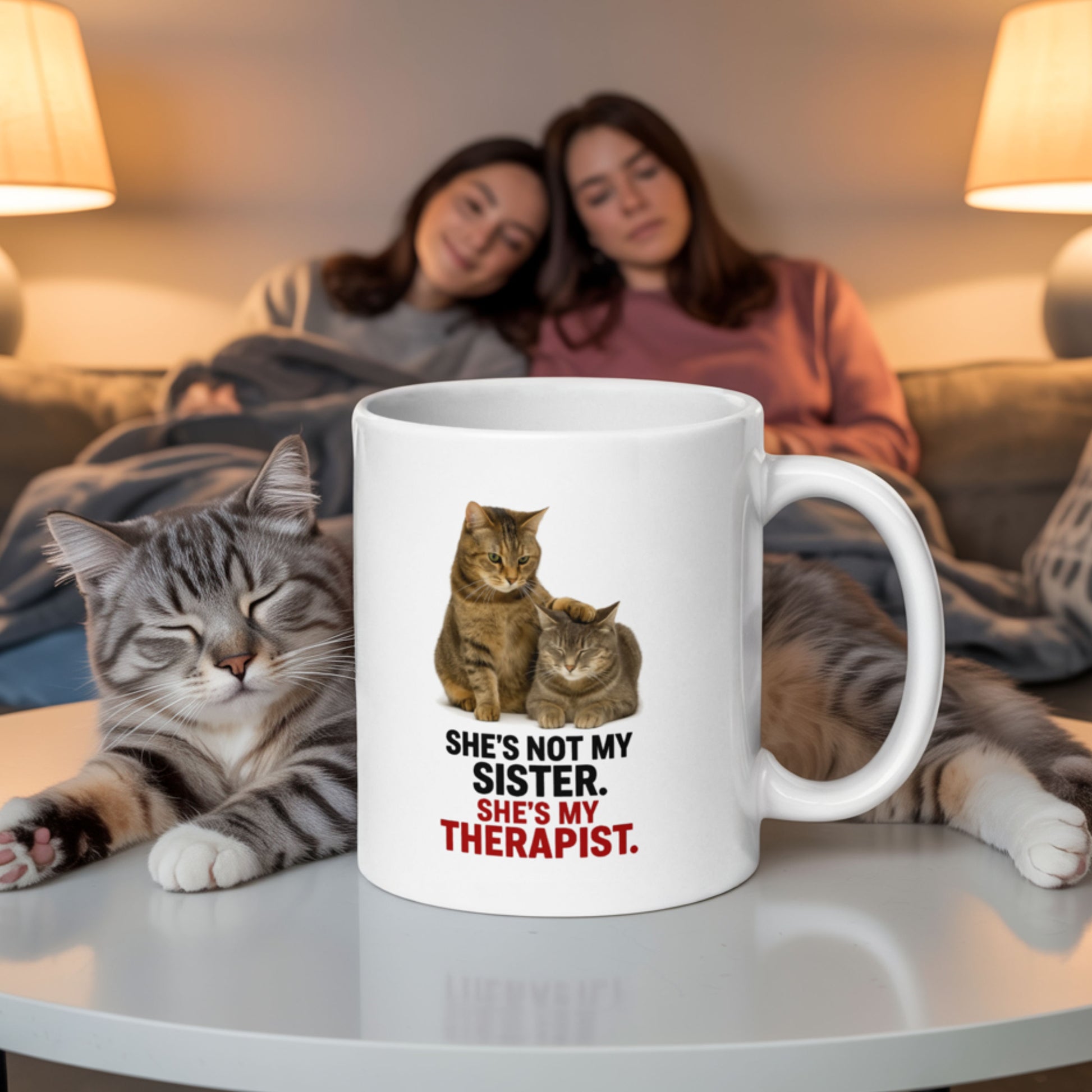 Funny sister mug that says 'She’s not my sister, she’s my therapist' featuring two cats, with one comforting the other, placed on a coffee table in front of two women relaxing on a couch and a real cat lounging nearby.